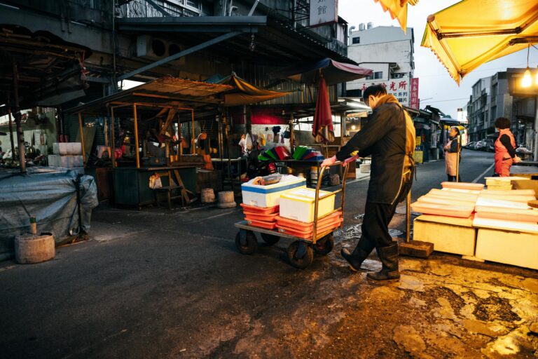 Busy city street market at dusk with a worker pushing a cart of boxes.